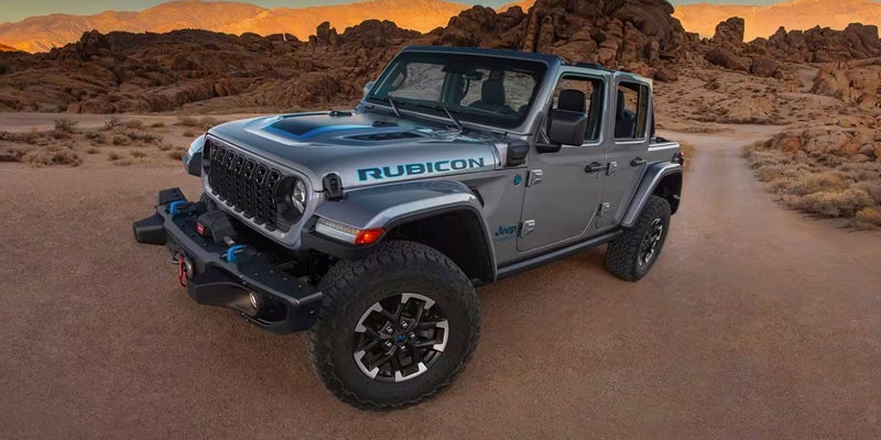 A grey Jeep Wrangler Rubicon 4xe on a dirt road in a desert landscape with rocky hills.