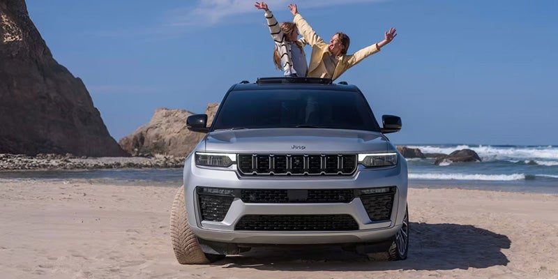 White Jeep Grand Cherokee SUV parked on a beach with passengers standing through the sunroof