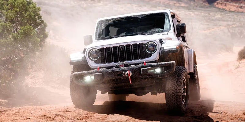 A white Jeep Wrangler Rubicon kicks up dust while off-roading on a dirt trail in a desert landscape